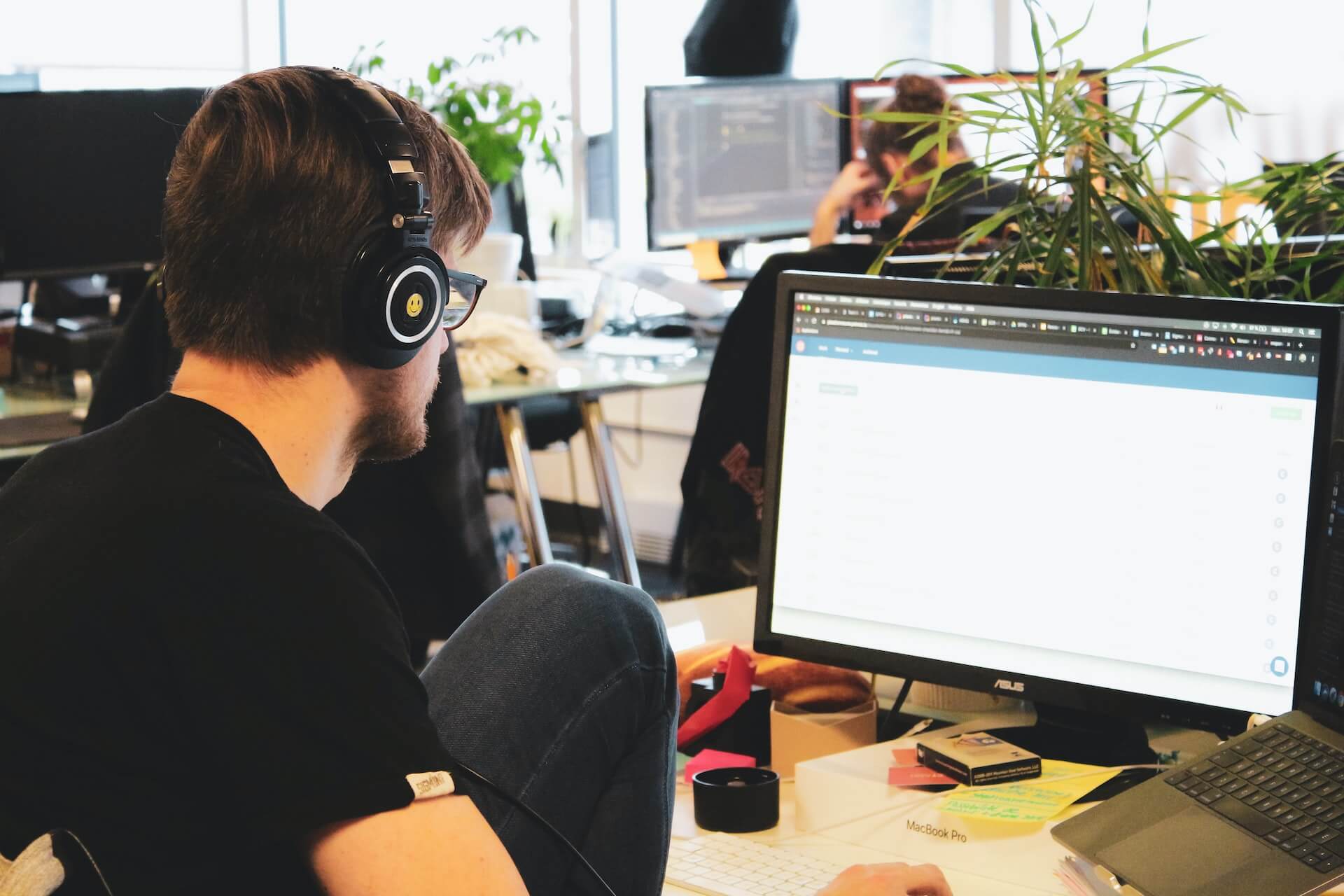 Young white man with headphones working at his computer. Other employees are in the background working at their desks in an office space.