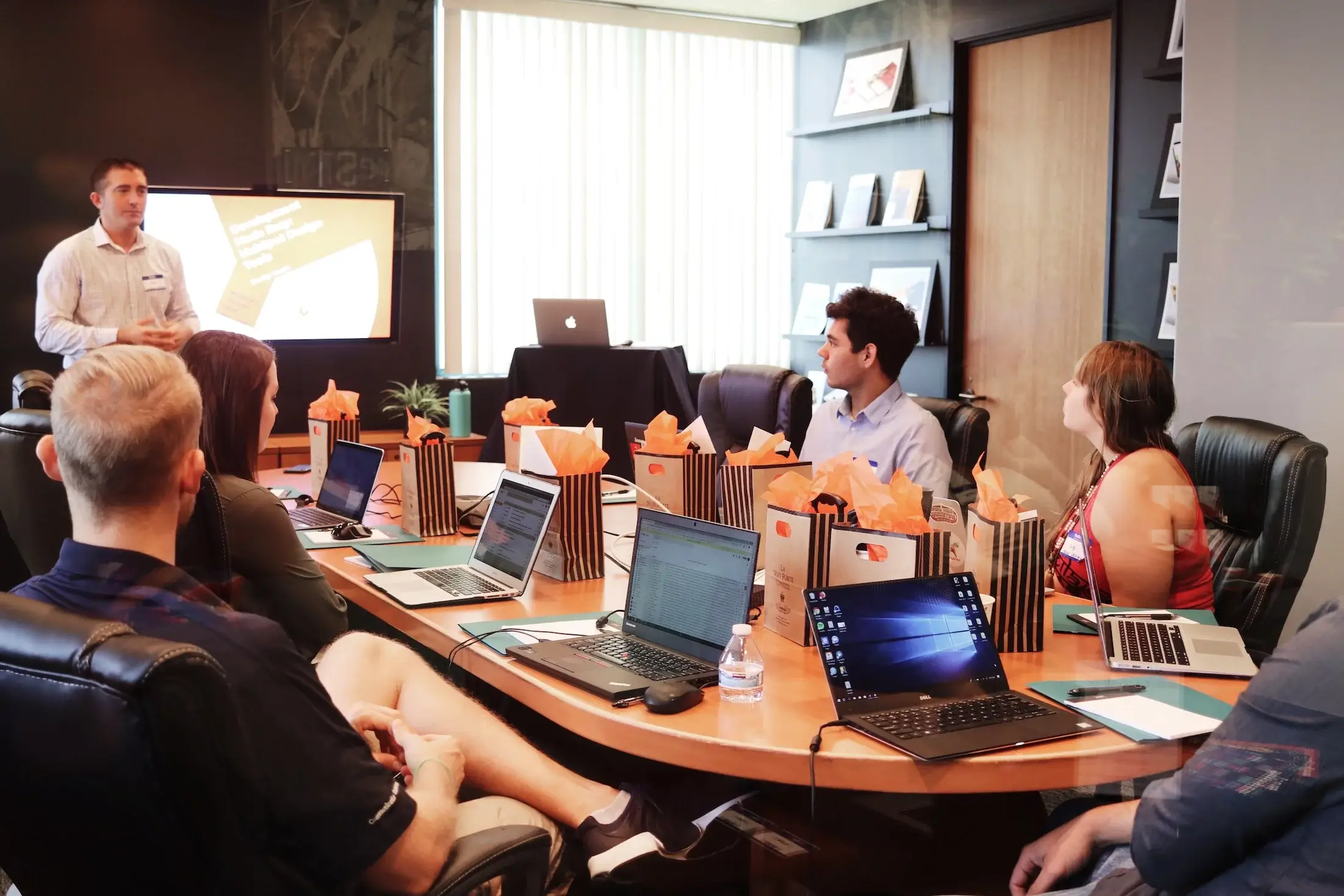 Small group of business people sitting around a conference table with one man standing up presenting with a projector screen